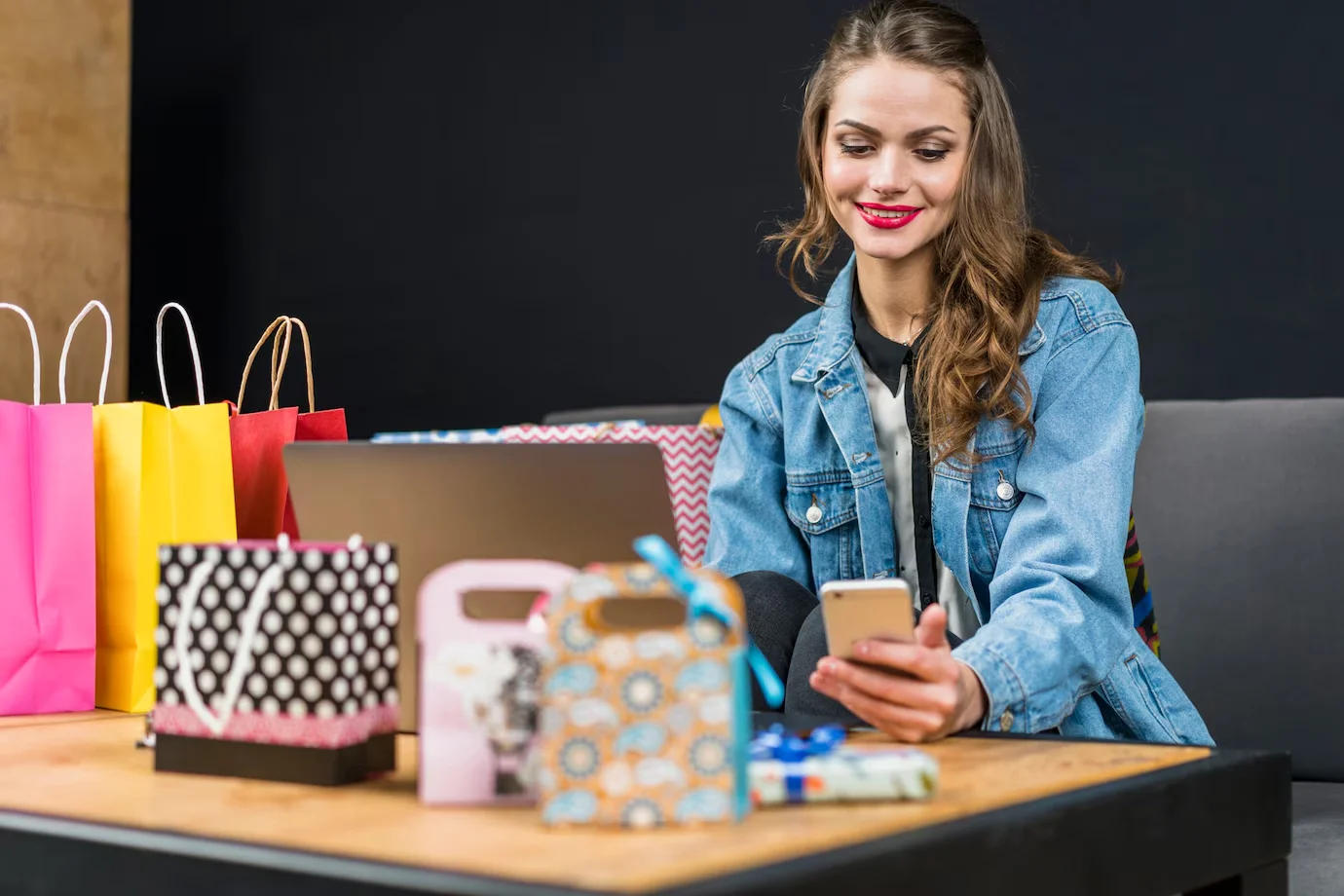 A stylish young woman wearing a denim jacket using her phone and laptop for online shopping.